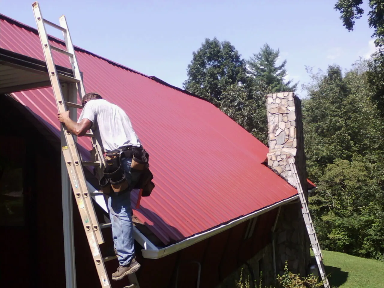 Roofer on a ladder beside a red metal roof and stone chimney — Soffit & Fascia Repair by Roofing Brothers in Pacific Palisades